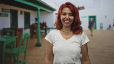 Terracotta fayansları olan bir binada, elleri yeşil sandalyelerin önünde gülümseyen ve barış işareti yapan bir kadın; neşe oyunculuğu doğallığı.