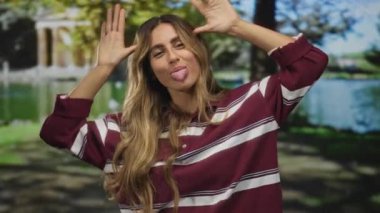 Güneşli sarı saçlı bir kadın yeşil bir parkta gülümserken başını eğiyor ve kollarını ardına kadar açıyor..