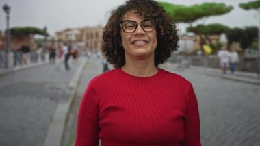 Kırmızı kazak giyen ve gözlüklü bir kadın parmaklarıyla barış işareti yapıyor ve sokakta gülümsüyor; neşeli iyimserlik.