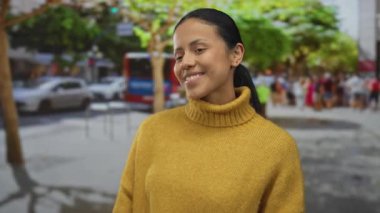 Kadın gülümsüyor ve hareketli caddede barış işareti yapıyor. Ağaçlar canlı şehir manzarası gösteriyor..