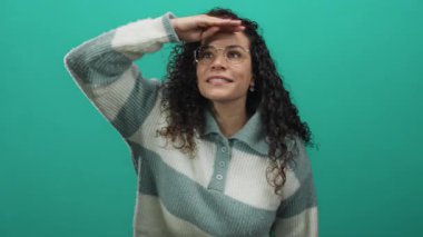 Gülümseyen ve yeşil arka planda ileriye bakan, gözlüklü ve çizgili süveter giyen genç İspanyol kadın, izole edilmiş canlı duvarın karşısında duruyor..