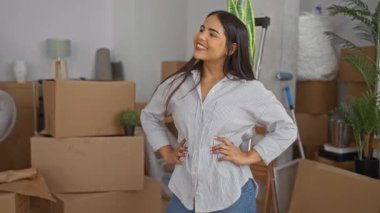 Yeni apartman dairesinde gülümseyen bir kadın. Etrafı kutu ve bitkilerle çevrili. Yeni bir taşınmaya işaret ediyor..