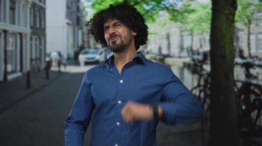 Elleri yüze yakın yürüyen ve kıvırcık saçlı bir adam, mavi gömlek görünür, park halindeki bisikletleri geçerken sırıtıyor;.