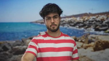 Sakallı, gözlüklü, çizgili tişörtlü genç adam kumlu sahilde, açık gökyüzünü işaret ederek kıyıda duruyor..