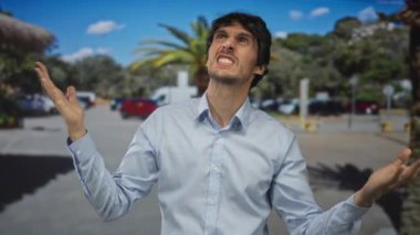 Açık mavi gömlekli genç adam güneşli bir şehir caddesinde el kaldırıyor ve kaşlarını çatıyor. Hayal kırıklığı..