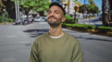 Arka planda ağaçlar ve binalar olan bir sokak ortasında gülümseyen İspanyol genç adam, güneşli bir günde rahat ve hoşnut bir havayı somutlaştırıyor..