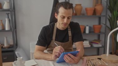 Stüdyoda Stylus ile tablet çizen adam, ekranda eller ve masada çömlek araçları; odaklanmış yaratıcılık.