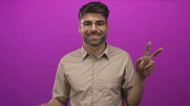 Adam mor stüdyoda iki eliyle barış işareti gösteriyor. Gözlük ve bej gömlek giyiyor. Zafer sevinci..