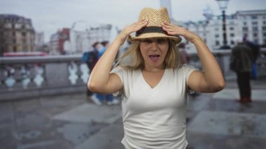 Kolları havada ve açık avuç içleri şehir köprüsünde hasır şapka ve beyaz t-shirt giyen, rüzgarla savrulan saçı olan bir kadın..