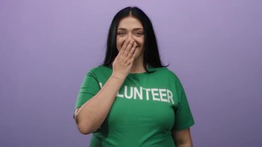Uzun saçlı, neşeli bir şekilde gülümseyen genç gönüllü kadın izole edilmiş mor arka plana karşı ağzını kapatıyor.