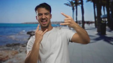 Genç İspanyol adam, açık havada mavi deniz ve palmiye ağaçlarıyla deniz kenarında coşkulu bir jest yapıyor.