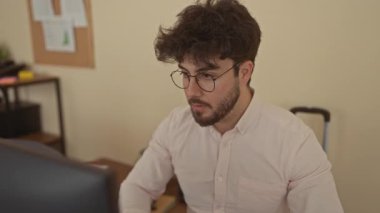 Ofisteki sakallı genç adam çenesini tutuyor, diş ağrısı çekiyor gibi görünüyor, kapalı alanda çalışıyor, modern çalışma alanındaki bilgisayara yoğunlaşırken rahatsızlık gösteriyor..