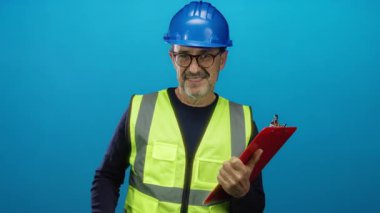 Emniyet yeleği ve kask giyen bir adam mavi arka planda kendine güvenen bir pano tutuyor. İş ortamında onay ve güvenceyi işaret ediyor.