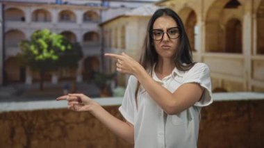 Kadın, gözlüklü ve beyaz gömlekli, çıplak kollu, eski bir kasaba sokağında sol tarafı işaret ediyor; şüphecilik kararsızlıktan şüpheleniyor..