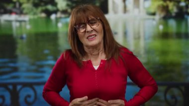 Gözlüklü, kırmızı kazaklı yaşlı bir kadın, arka planda yemyeşil bir gölle manzaralı bir parkta dikilirken acı çekiyor.