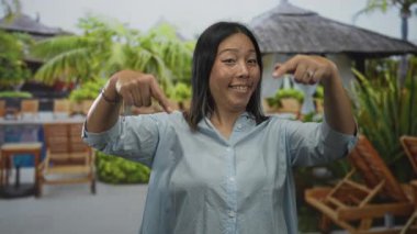 Kabin ve şezlongun yanındaki havuz kenarını gösteren kadın, gömlek ve bilezik takıyor; dostça bir davet..