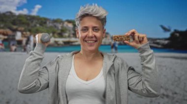 Kısa gri saçlı kadın bir elinde 1 kg ağırlığında bir dambıl ve stüdyo sahnesinde yüzüne yakın bir granola barı tutuyor..