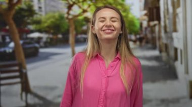 Canlı bir şehir caddesinde neşeli bir şekilde gülümseyen kadın, canlı pembe bir gömlek giyiyor ve arka planda ağaçlar var. Açık hava manzarası yaratıyor..