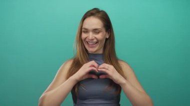 Tebessüm eden beyaz bir kadın parlak deniz mavisi stüdyosunda elleri ile kalp hareketi yapar..