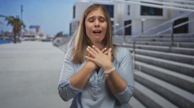 Sarışın genç bir kadın, kentin dış sokağında beton merdivenlerle ve modern bina cephesiyle ağrı için göğüs debriyajı kullanıyor..