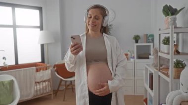 Hamile kadın kulaklıkla müzik dinliyor ve parlak bebek odasında telefon kullanıyor, oyuncaklarla dolu beşik ve rafların yanında gülümsüyor, rahat ve modern bir ev ortamı sergiliyor..