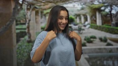 Genç İspanyol kadın, mavi tişört giyen, taş sütunlar ve yeşil bitkilerle birlikte avluda başparmak göğse işaret ediyor..