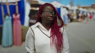 Açık havada, örgü ve gözlüklü bir kadın renkli giyim mağazalarının yanındaki bir markette gün boyunca hoşnutsuz görünüyor..