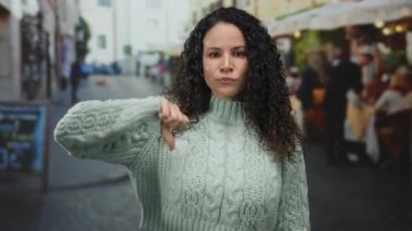 Dışarıdaki terasta kıvırcık saçlı, süveter giyen, kafelerle çevrili sokakta duygularını ifade eden ve gündüzleri arka planda olan kadınlar..