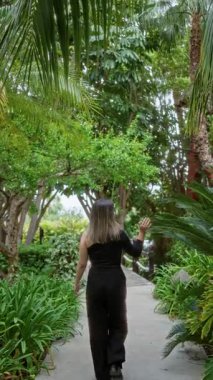 Kadın bereketli bir tropikal otel yolunda yürüyor. Etrafı canlı yeşilliklerle çevrili, huzurlu bir tatil köyünde. Palmiye yapraklarından bir gölgeliğin altında..