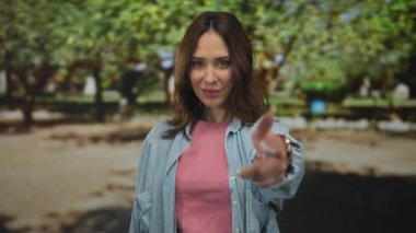 Pembe gömlekli ve kot ceketli genç İspanyol kadın güneşli bir parkta gülümseyip kamerayı işaret ederek yeşil ağaçlarla neşeli ve çekici bir açık hava yaratıyor..