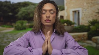 Mor gömlekli kadın kulaklık takıyor ve avuçlarını yemyeşil parkta birbirine bastırarak gözlerini kapıyor..