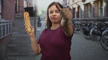 Öndeki bir şehir caddesindeki kameraya başparmağıyla bir baget tutan genç kadın; şehir onaylamaması.