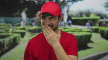 Kırmızı şapkalı ve gömlekli adam, sokak parkında ağzı ve yeşil çitleri güneş ışığı altında kaplayan; utangaç eğlence.