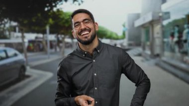 Şehir sokaklarında gülümseyen ve düşünen genç bir adam, gözlüklü ve sakallı sıradan bir stil sergiliyor, canlı bir kasaba atmosferini gösteren bir şehrin arka planına kurulmuş..