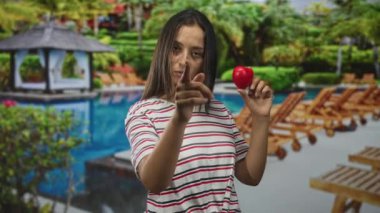 Çizgili gömlekli kadın elinde küçük kırmızı bir kalp tutuyor ve diğer elini havuz kenarındaki çardak ve şezlonglarda kameraya doğru uzatıyor..