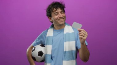 İspanyol bir adam elinde mor bir arka planda futbol topu ve kart tutuyordu. Mavi gömlek ve atkı takmış gülümsüyordu. Neşeli spor coşkusunu somutlaştırıyordu..