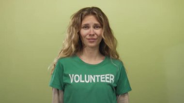 Stüdyo ortamında, sarışın gönüllü bir tişörtle genç bir sırıtışı sergileyen yeşil bir duvara karşı ezik tabelası oluşturan bir kadın..