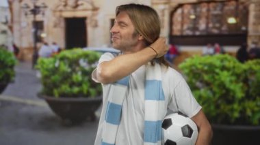 Uzun saçlı, çizgili atkı takan sarışın bir adam futbol topunu kalabalık bir sokakta tutuyor..