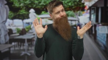 Uzun kızıl sakallı bir adam gün ışığında restoran terasında avuçlarını kaldırıyor; belirsizlik karışıklığı.