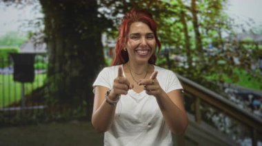 Kadın, gülümseyen ve öne doğru eğilen bir orman parkında çıplak elle işaret ediyor; neşeli bir teşvik..