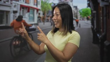 İşaret parmaklarıyla işaret eden bir kadın, alışveriş merkezleri ve bisikletlerin yakınındaki bir caddede gülümserken, neşeli bir merak.