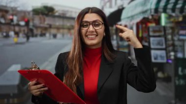 Gözlüklü genç bir kadın işlek caddede kırmızı pano tutarken gülümsüyor ve bulutlu gökyüzünün altında kafasını işaret ederek canlı şehir enerjisi ve özgüvenini yakalıyor.