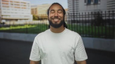 Beyaz tişörtlü bir adam binanın önünde yumruk yumruğa gülümseyip öpücük savuruyor, sakal ve kollarını gösteriyor, sevgi dolu bir teklif..