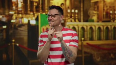 Çizgili tişörtlü genç adam gözlüklü ve dövmeli bir kilisenin içinde gülümsüyor, neşeli ifadesini ve renkli geçmişini vurguluyor..