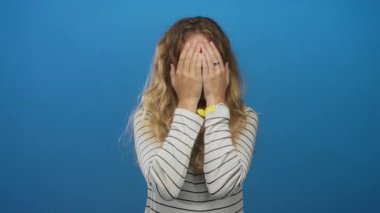 Genç bir kadın bir stüdyo sahnesinde ellerini sürpriz bir şekilde tutuyor. Mavi bir arka planda sarı bukleli, etkileyici bir portrede gülümsemesi var..