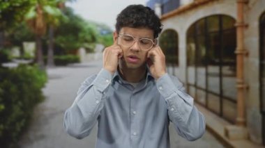 İnsanlar, ağaçlarla ve yeşilliklerle dolu güneşli bir sokakta dikilirken kulaklarını parmaklarıyla kapatırlar..