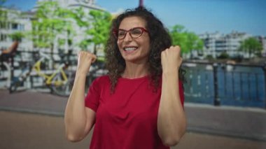 Kırmızı gömlekli ve kırmızı gözlüklü bir kadın kaldırılmış yumruklarıyla kanal boyunca gülümseyen kıvırcık saçlı bir kadın, zafer sevinci..