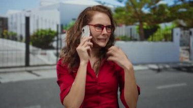 Kırmızı gözlüklü, kırmızı bluzlu kadın güneşli şehir caddesinde telefonda heyecanlı bir şekilde konuşuyor..