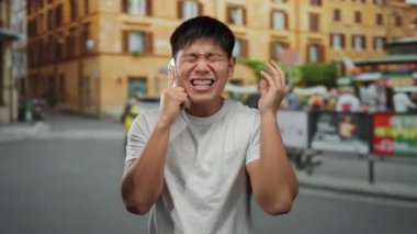 Cep telefonuyla konuşan genç adam şehir caddesinde hayal kırıklığına uğramış görünüyor. Mimarisi bulanık ve arka planda insanlar var..