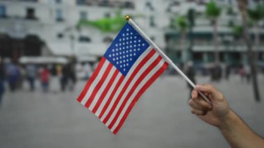 Kalabalık bir sokakta Amerikan bayrağını tutan adam, bulanık şehir arka planında canlı renkler ve şehir sahnesinde çeşitli hareketlerle vatanseverlik temasını vurguluyor..